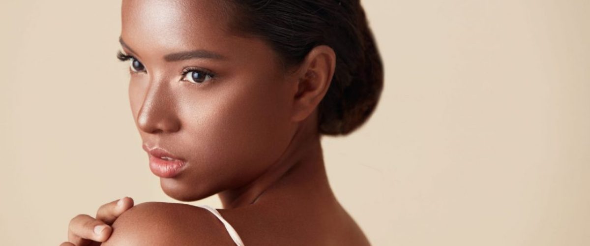 Beauty. Mixed Race Woman Portrait. Beautiful Ethnic Model Touches Her Shoulder And Looking Away. Female With Perfect Glowing Skin And Nude Natural Makeup Posing Against Beige Background.